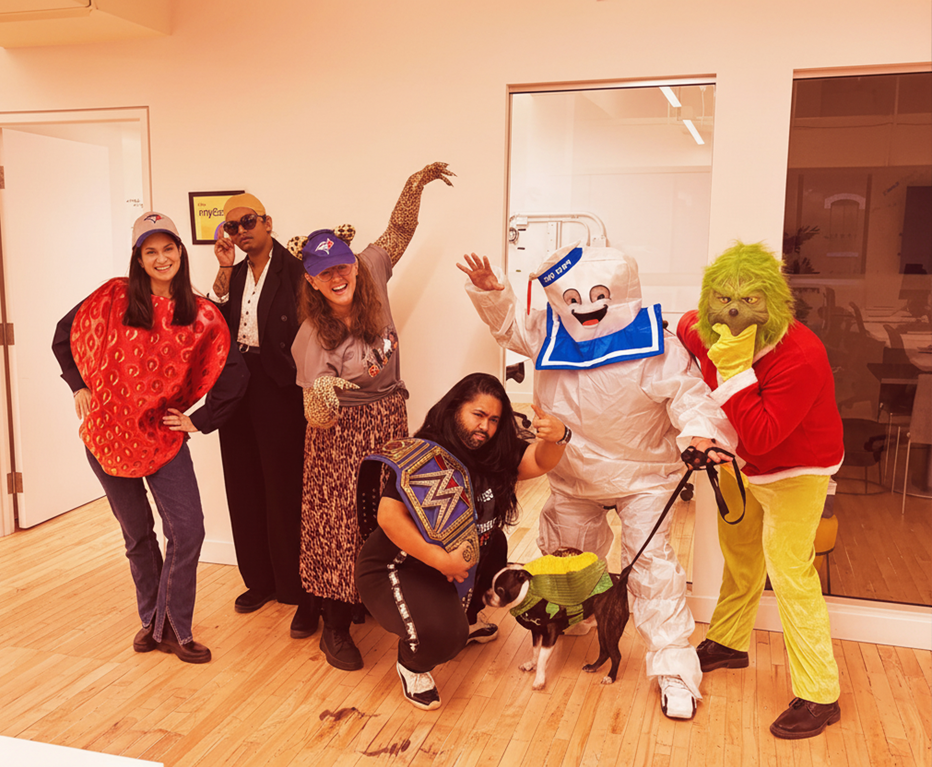 Six adults and a dog in a room dressed in various costumes including a strawberry, leopard print, wrestling champion, Stay Puft Marshmallow Man, and the Grinch, with the dog wearing a corn costume.