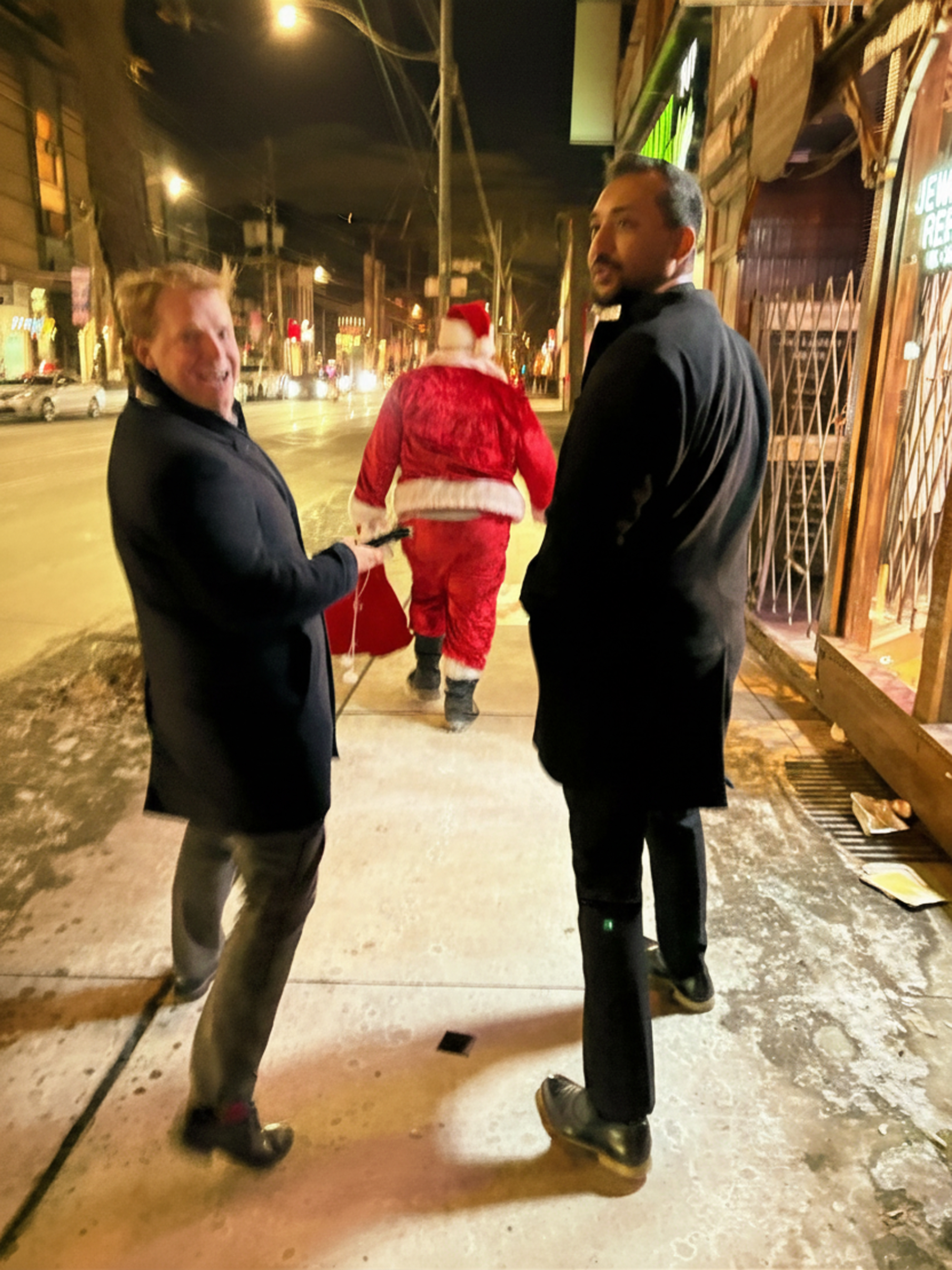 Two men in black coats walk on a sidewalk at night behind a person dressed in a Santa Claus costume.
