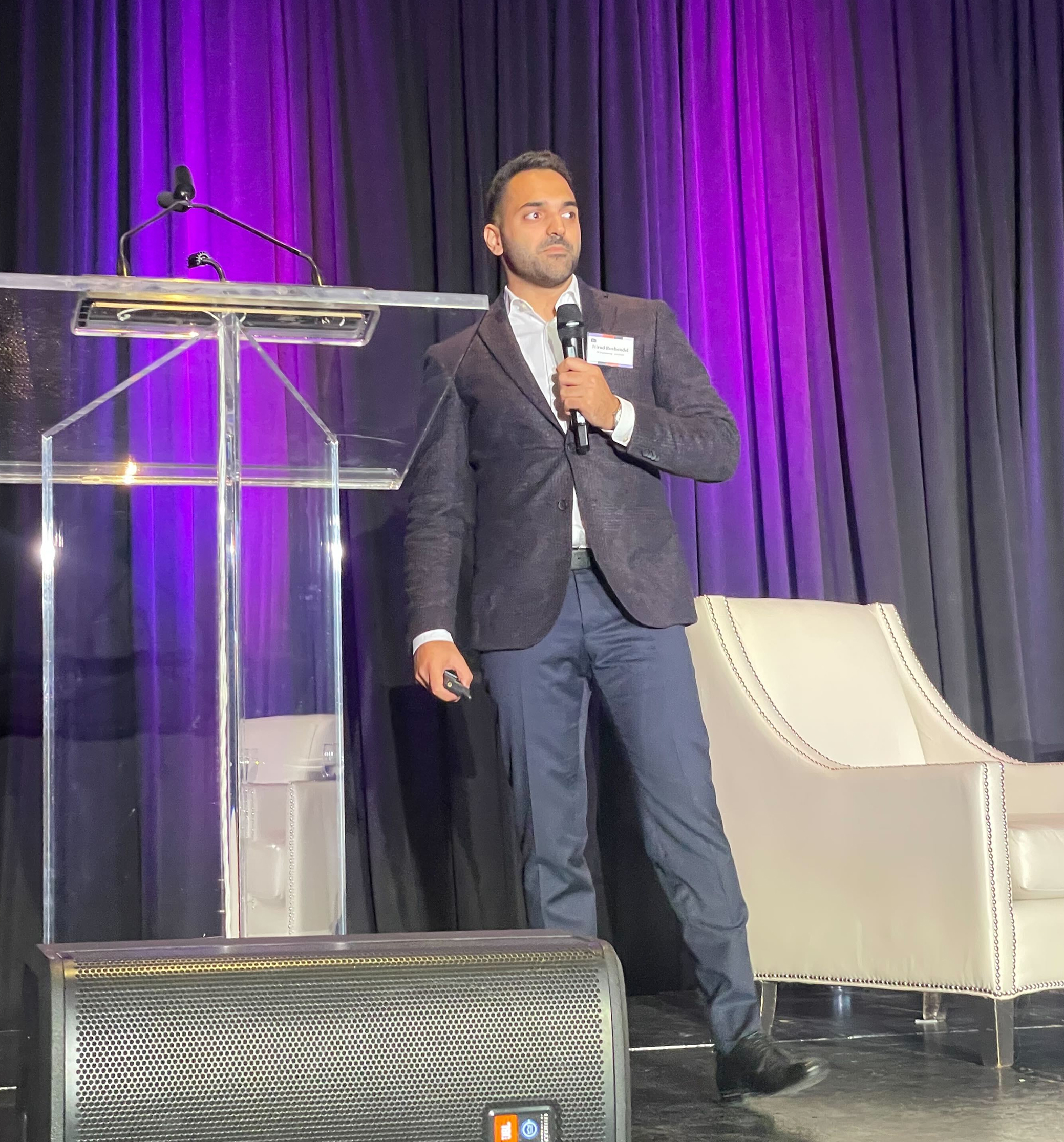 Man in a dark blazer speaking into a microphone on stage next to a clear podium and beige armchair, with purple curtains in the background.