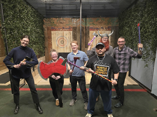 Man in a dark blazer speaking into a microphone on stage next to a clear podium and beige armchair, with purple curtains in the background.