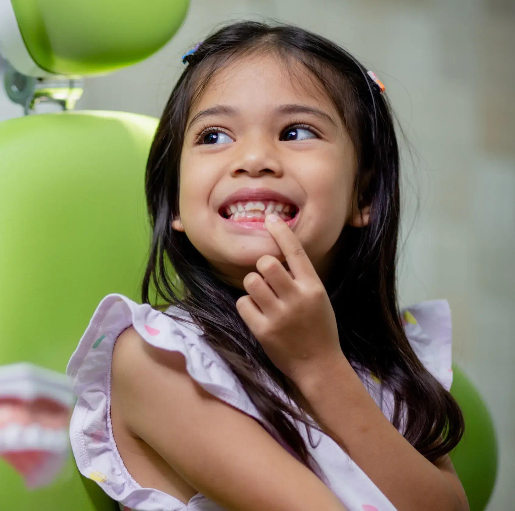 Smiling young girl pointing to her teeth while sitting in a green dental chair.
