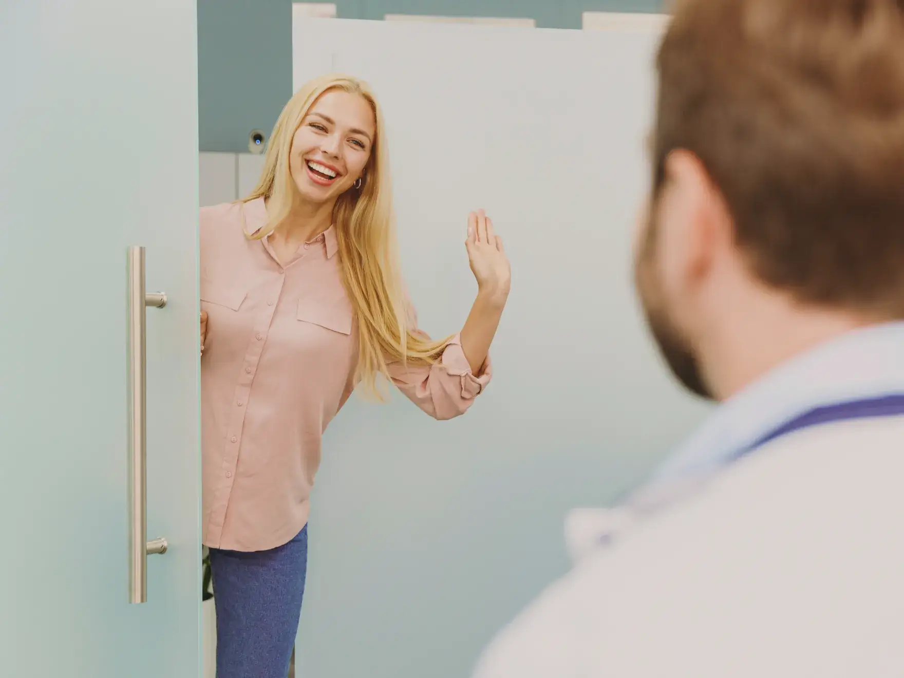 Smiling woman waving while standing at a partially open door, facing a man.
