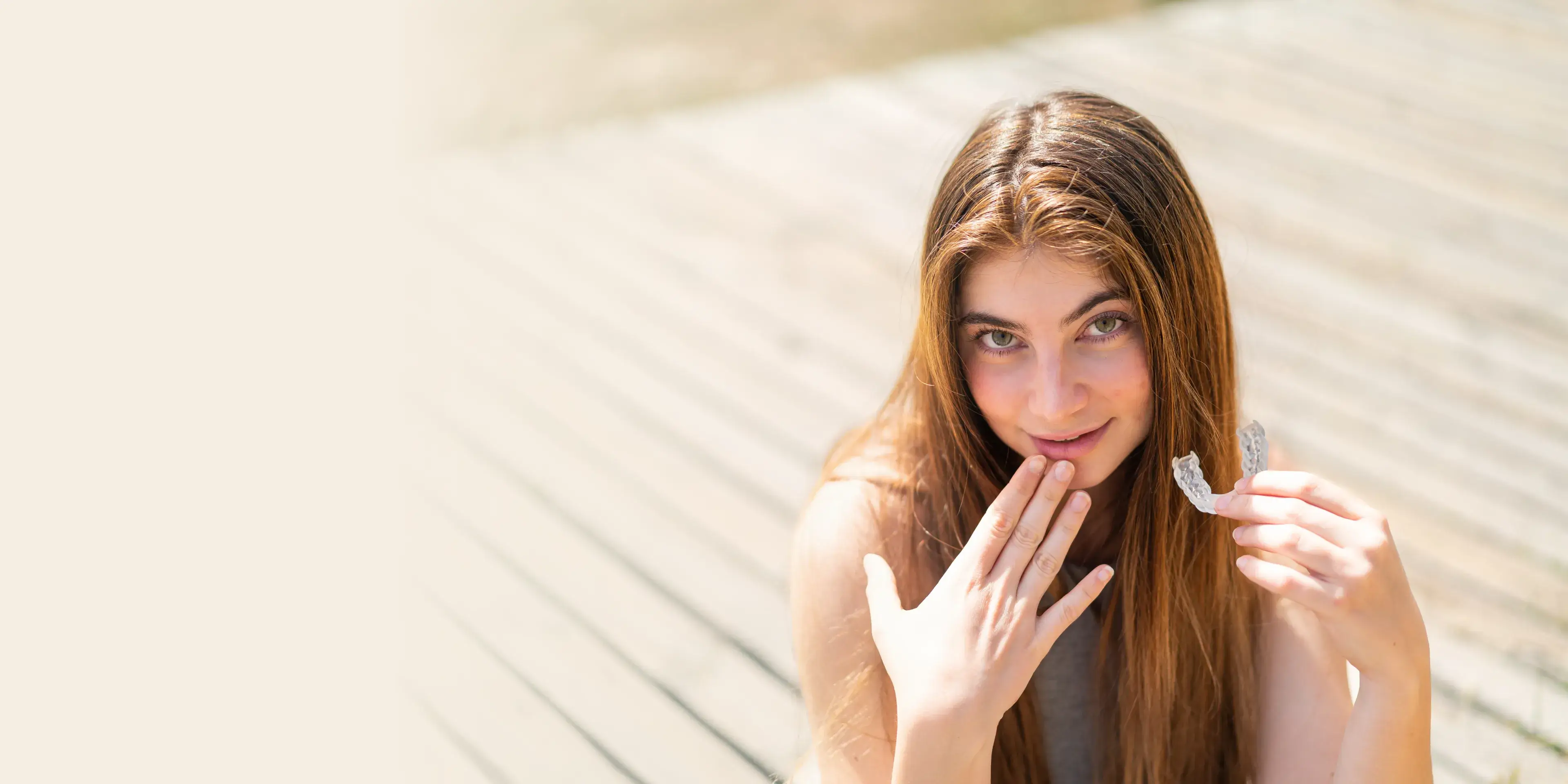 Young woman with long brown hair holding a clear dental aligner and smiling outdoors.