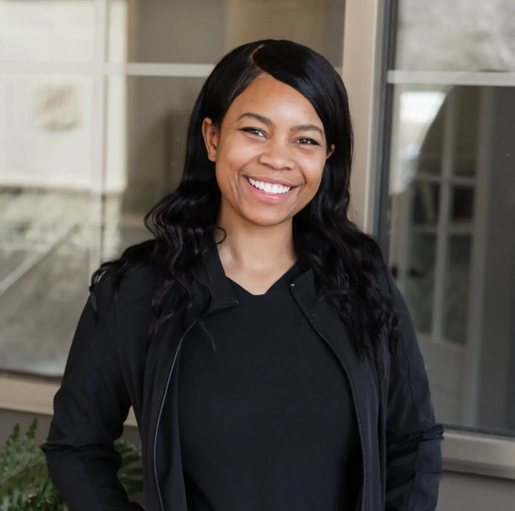 Smiling woman with long black hair wearing a black zip-up jacket standing indoors.