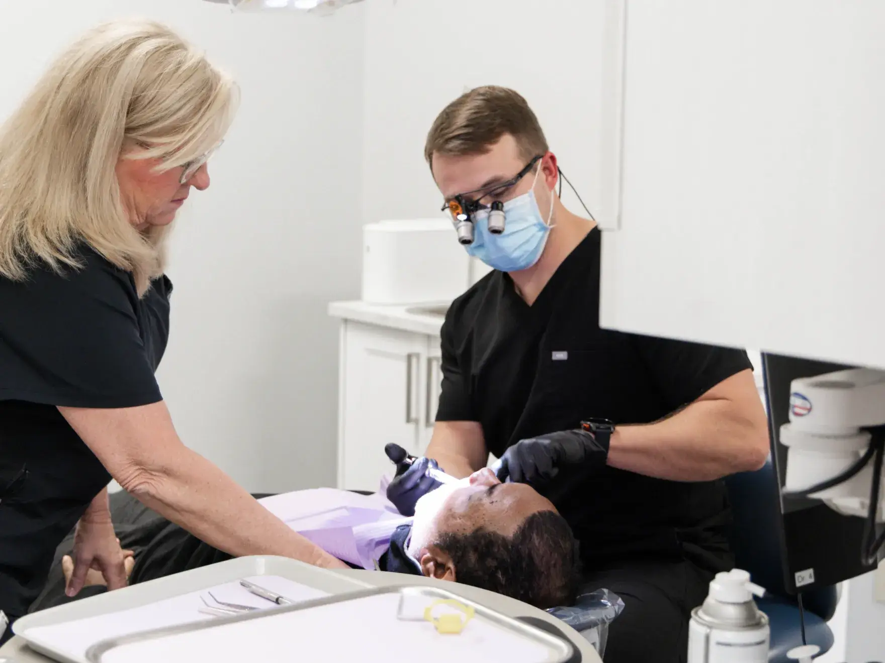 Dentist wearing magnifying glasses and mask treating a patient lying in a dental chair with an assistant nearby.