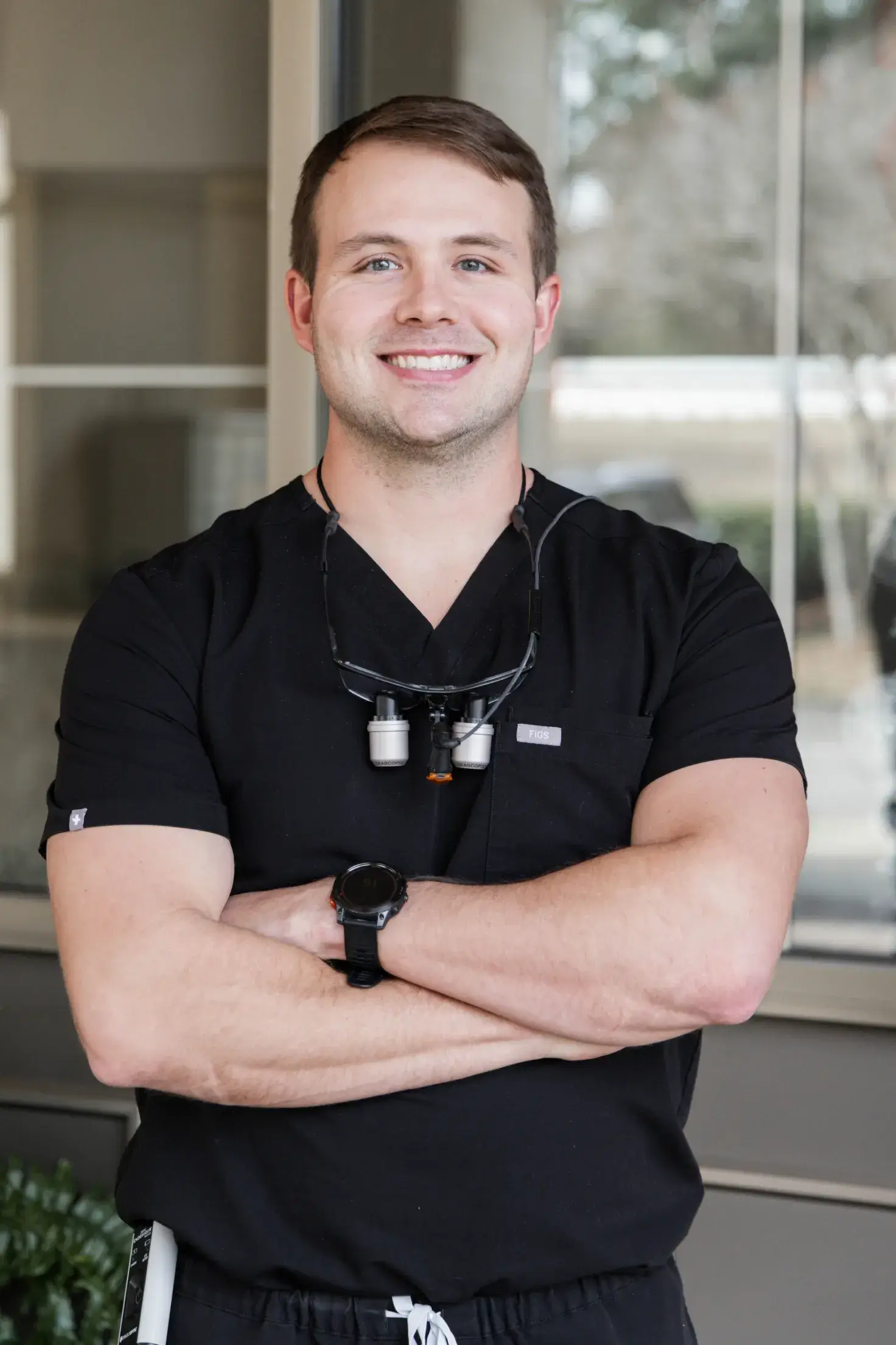 Smiling male healthcare professional in black scrubs with folded arms and dental loupes around his neck.