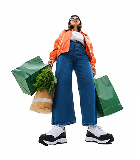 Low-angle view of a person wearing an orange jacket and wide jeans holding green shopping bags with groceries.