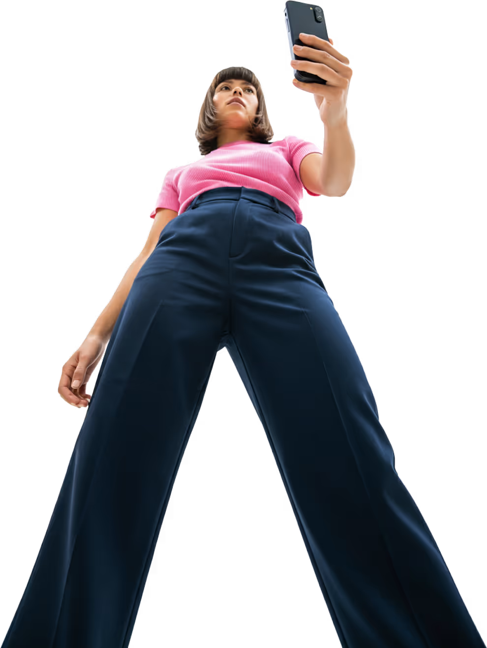 Woman in a pink shirt and blue wide-leg pants taking a selfie with her smartphone from a low angle.