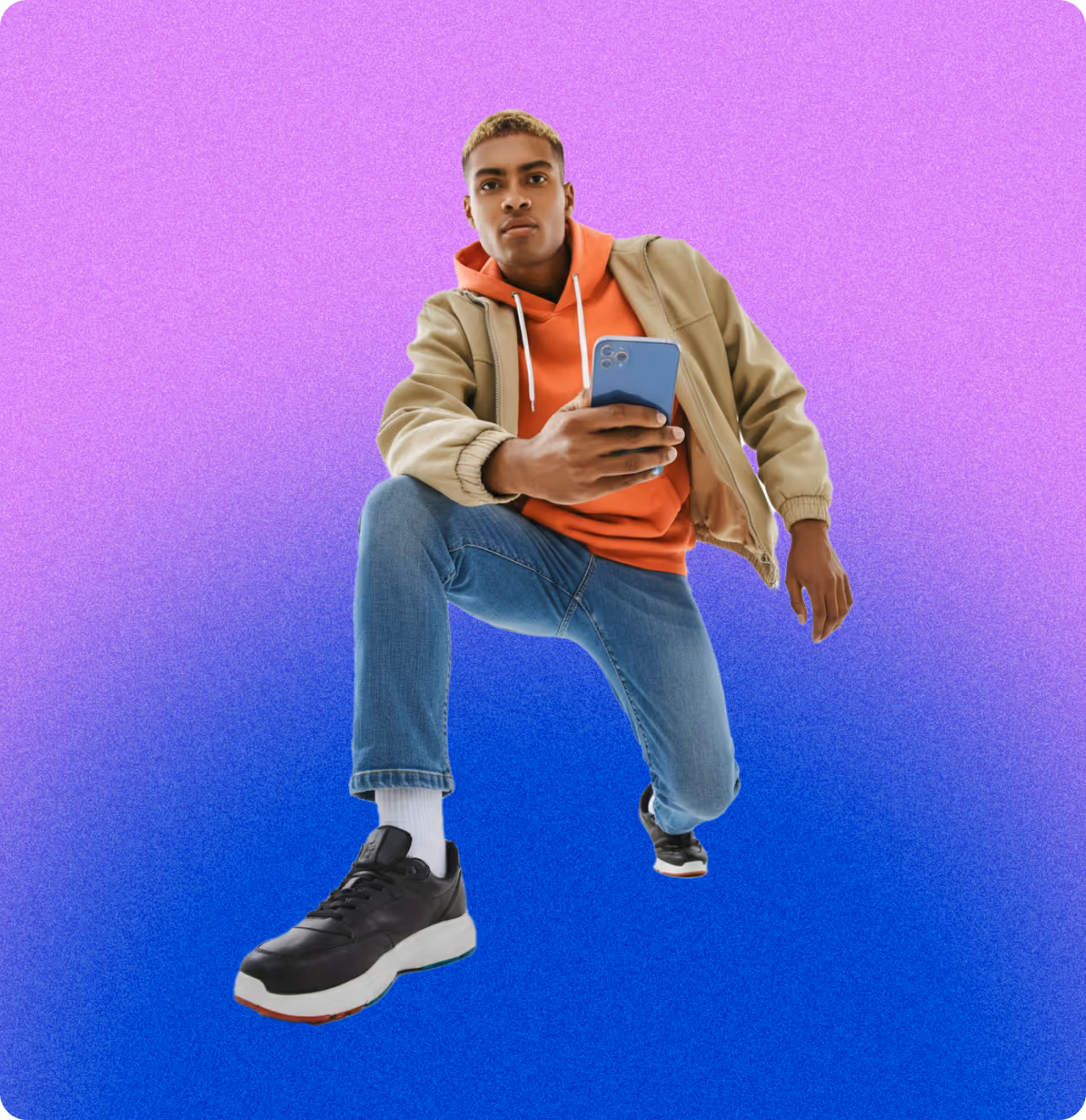 Young man with blonde hair wearing an orange hoodie, beige jacket, jeans, and black sneakers, holding a smartphone against a purple and blue gradient background.