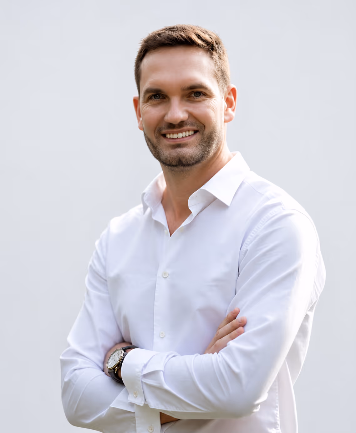 Smiling man with short hair in a white button-up shirt with arms crossed against a plain background.