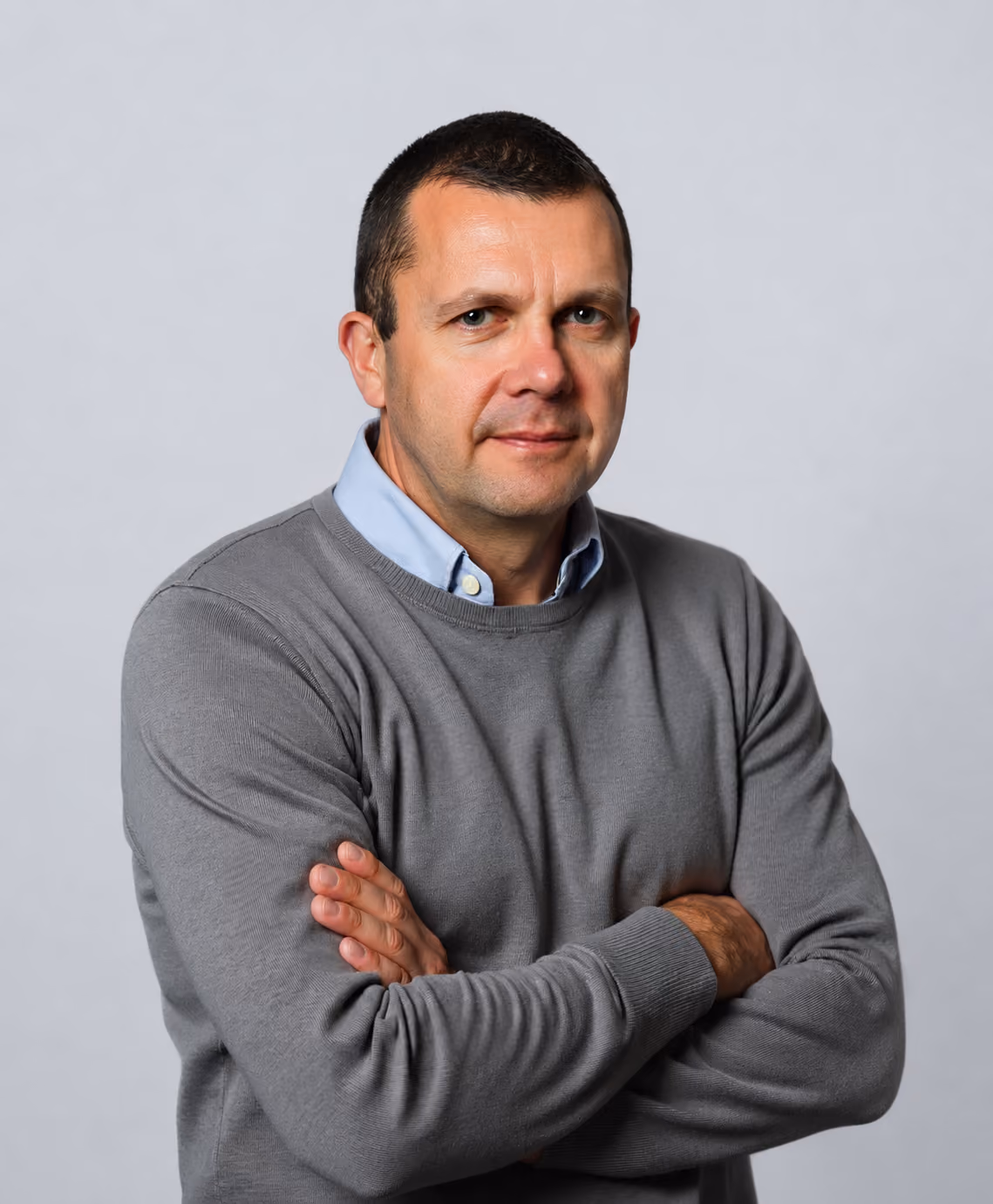 Man with short hair wearing a gray sweater over a blue collared shirt, arms crossed, against a gray background.