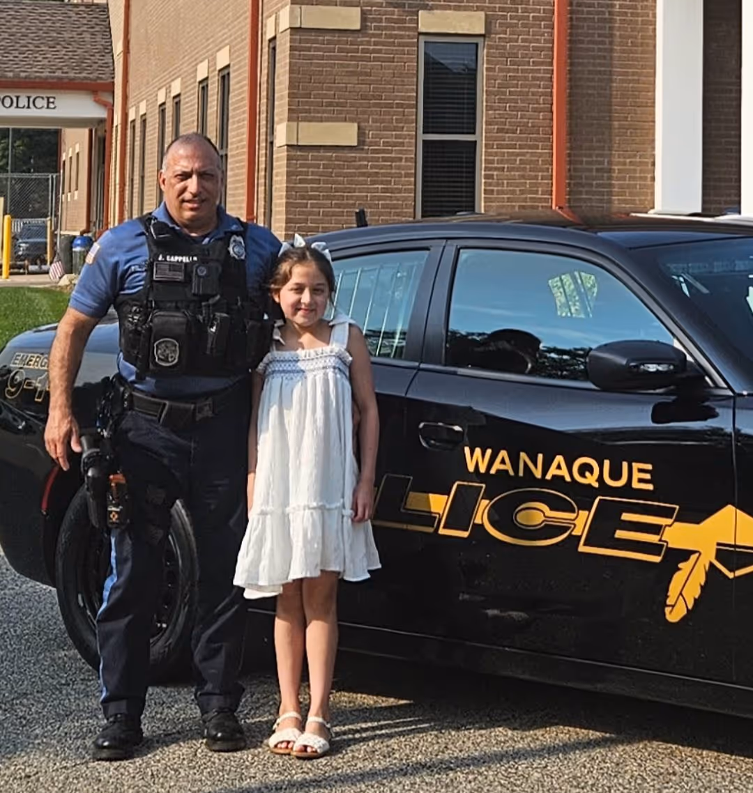Police officer and young girl standing together beside a Wanaque Police car in front of a brick building.