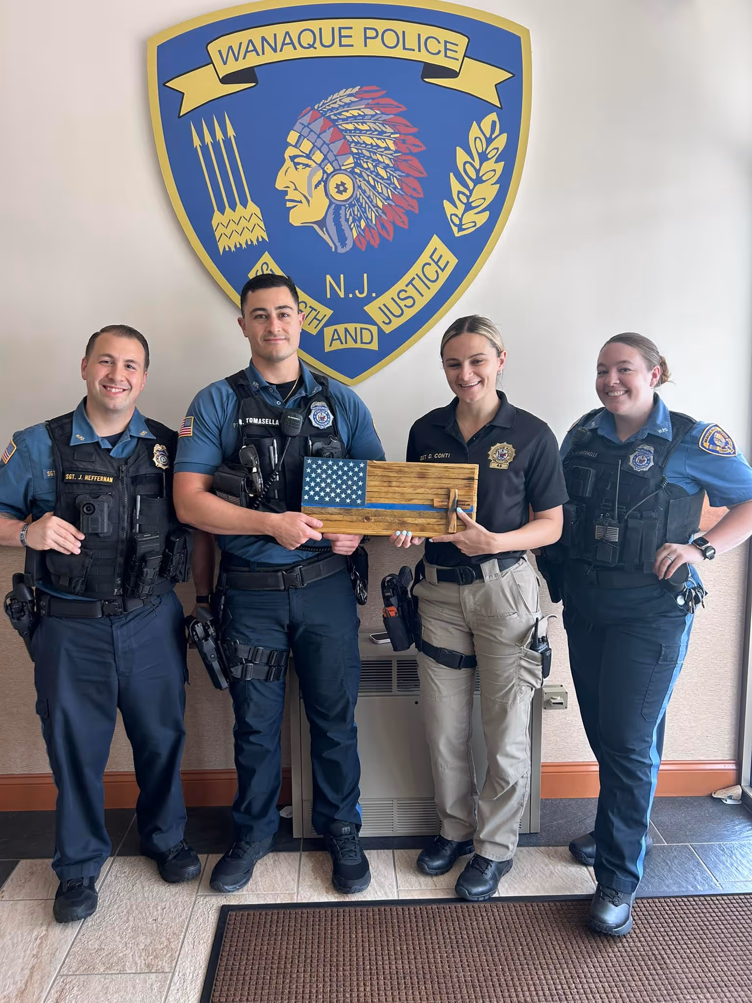 Four Wanaque Police officers smiling and holding a wooden American flag plaque beneath the Wanaque Police emblem on a wall.