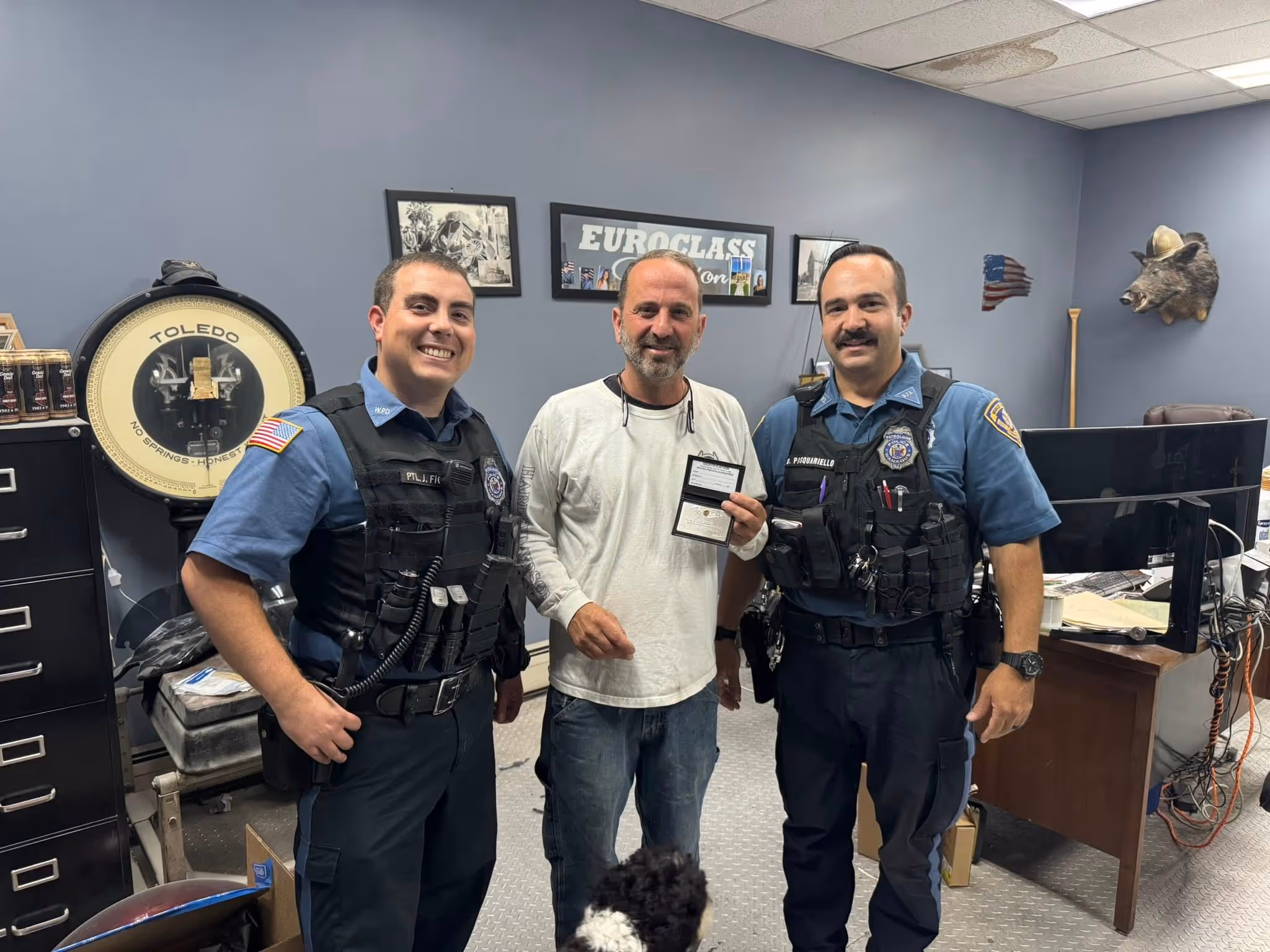 Two uniformed police officers standing on either side of a man holding an ID badge inside an office.