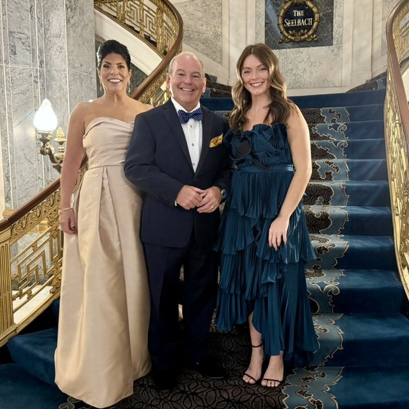 Two women and one man dressed in formal attire standing on a grand staircase with ornate gold railings and a blue carpet at charity benefit.