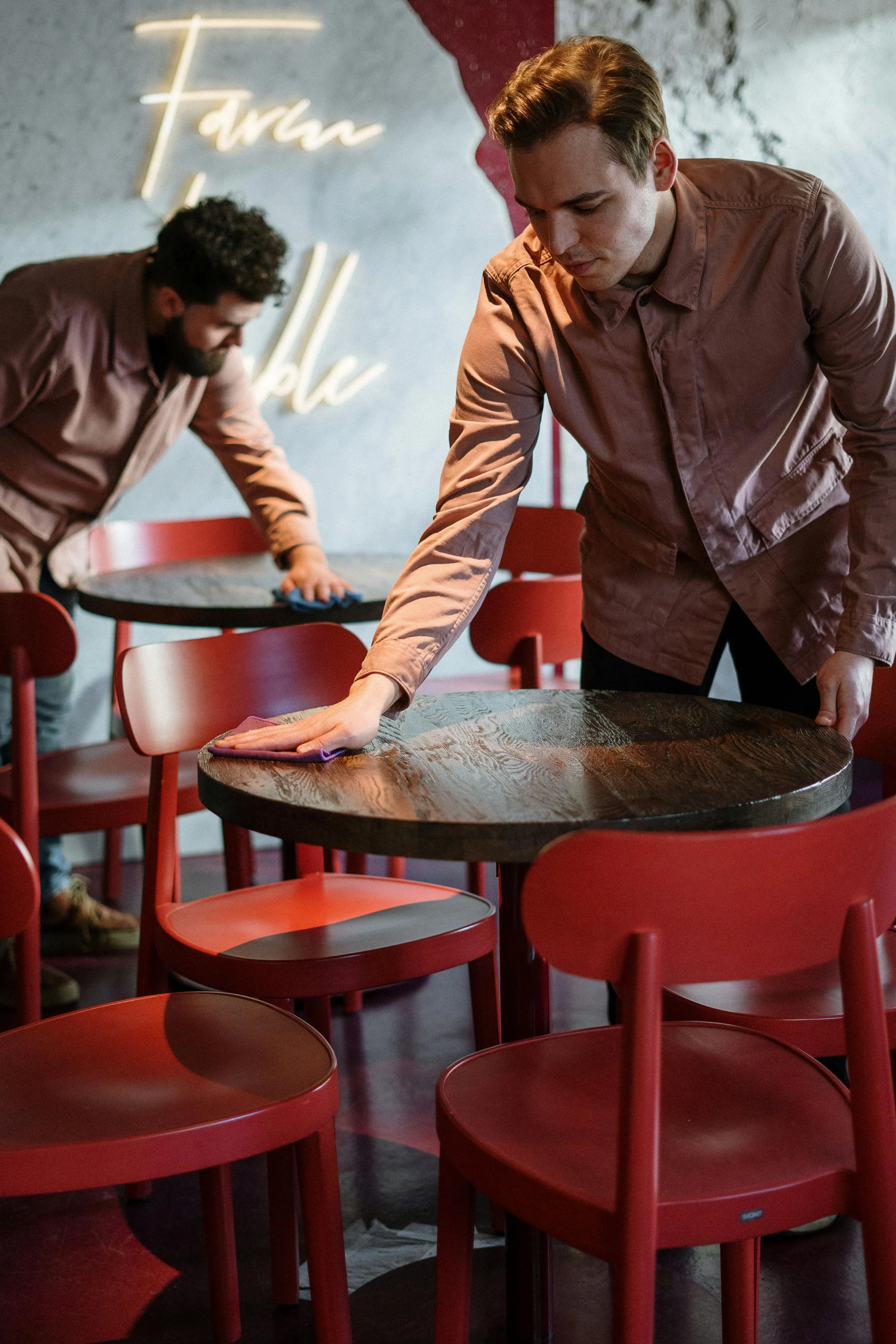 Two men in brown jackets cleaning wooden tables in a restaurant with red chairs.