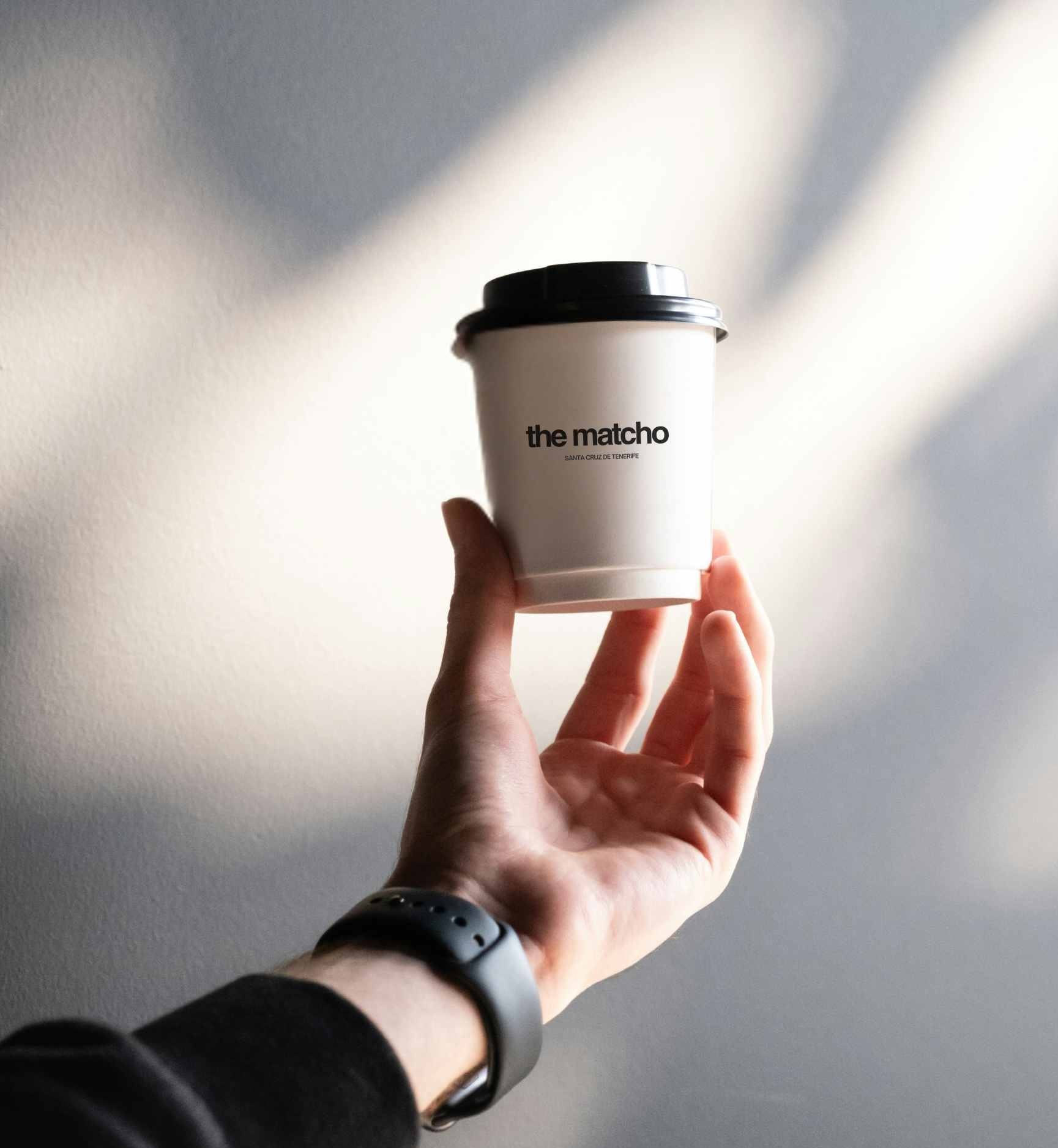Hand holding a white coffee cup with black lid labeled 'the matcho' against a softly lit wall.