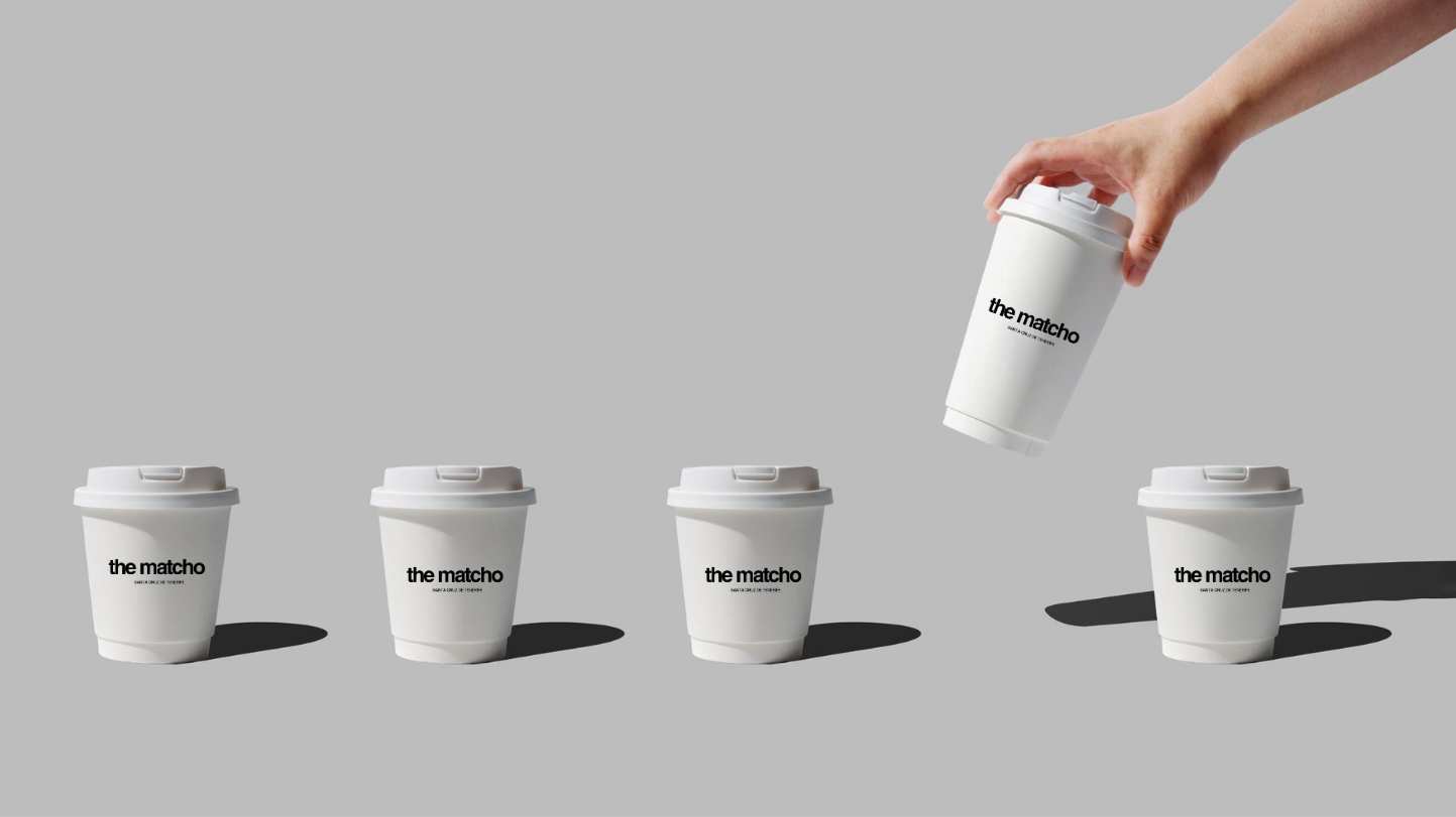 A hand holding a white disposable coffee cup tilted above three identical cups standing on a gray surface, all labeled 'the matcho'.