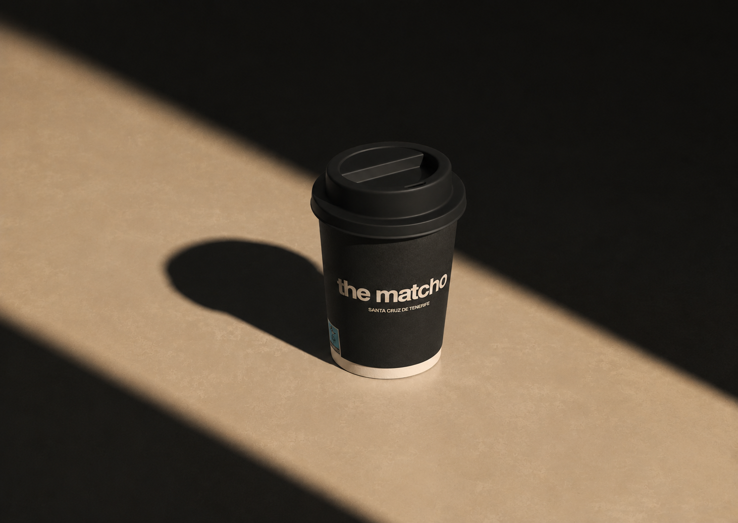 Black takeaway coffee cup with lid labeled 'the matcho Santa Cruz de Tenerife' in sunlight and shadow.