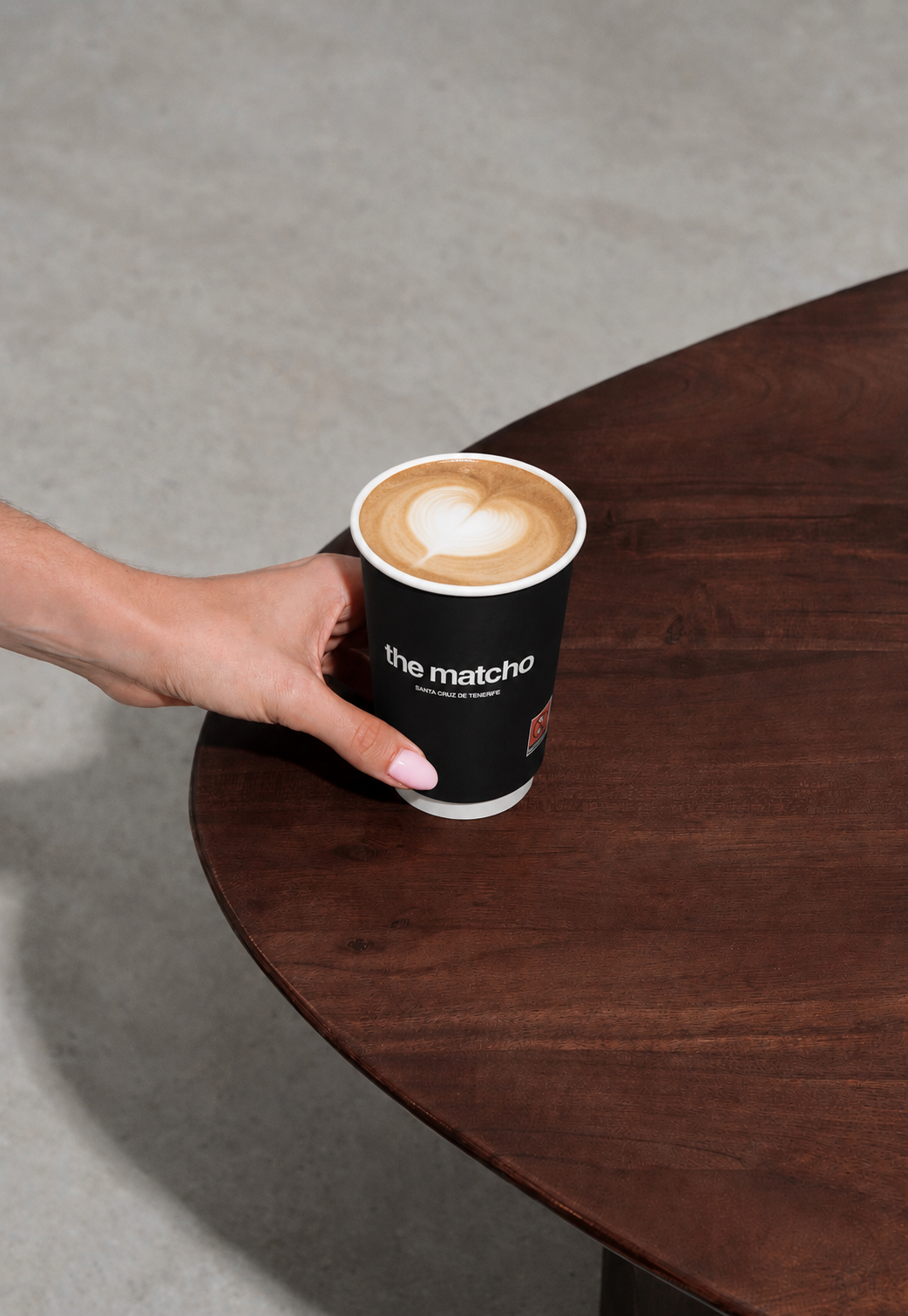 Hand lifting a plastic lid off a cup with green liquid splashing inside on a textured pavement.