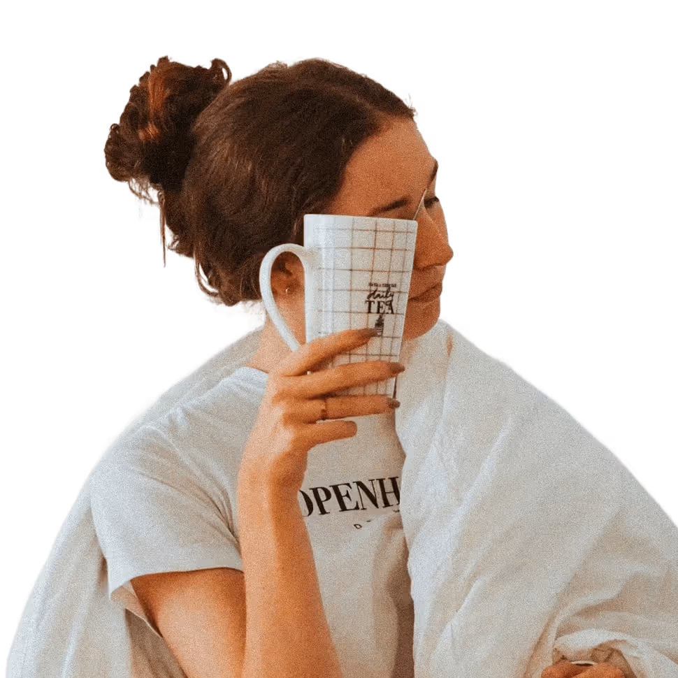 Woman with brown hair in a bun, wrapped in a white blanket, holding a white grid-patterned mug close to her face.