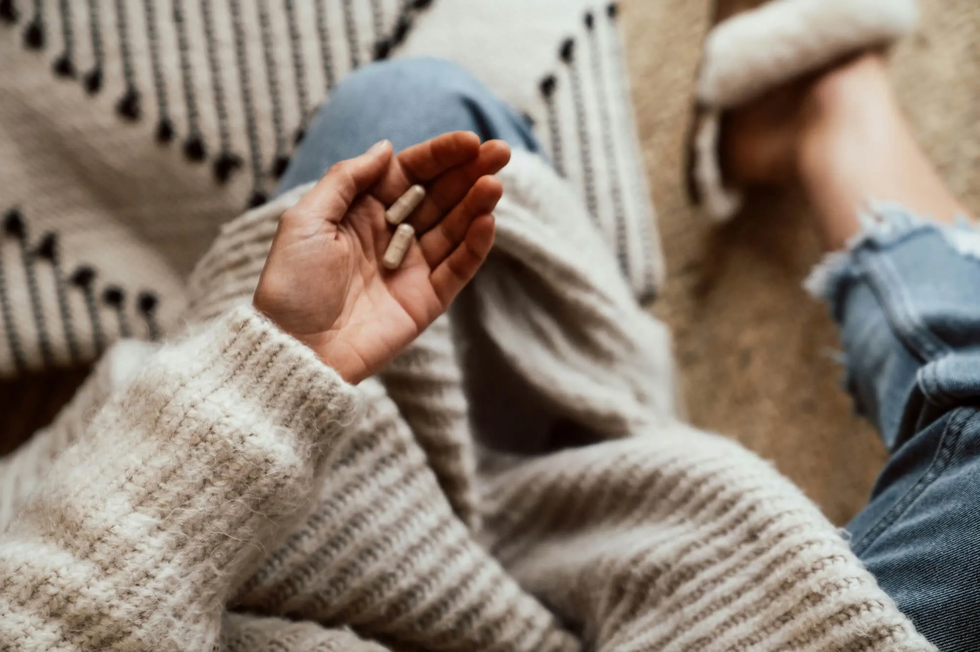 Person sitting cross-legged holding two white capsules in hand, wearing cozy sweater and jeans.
