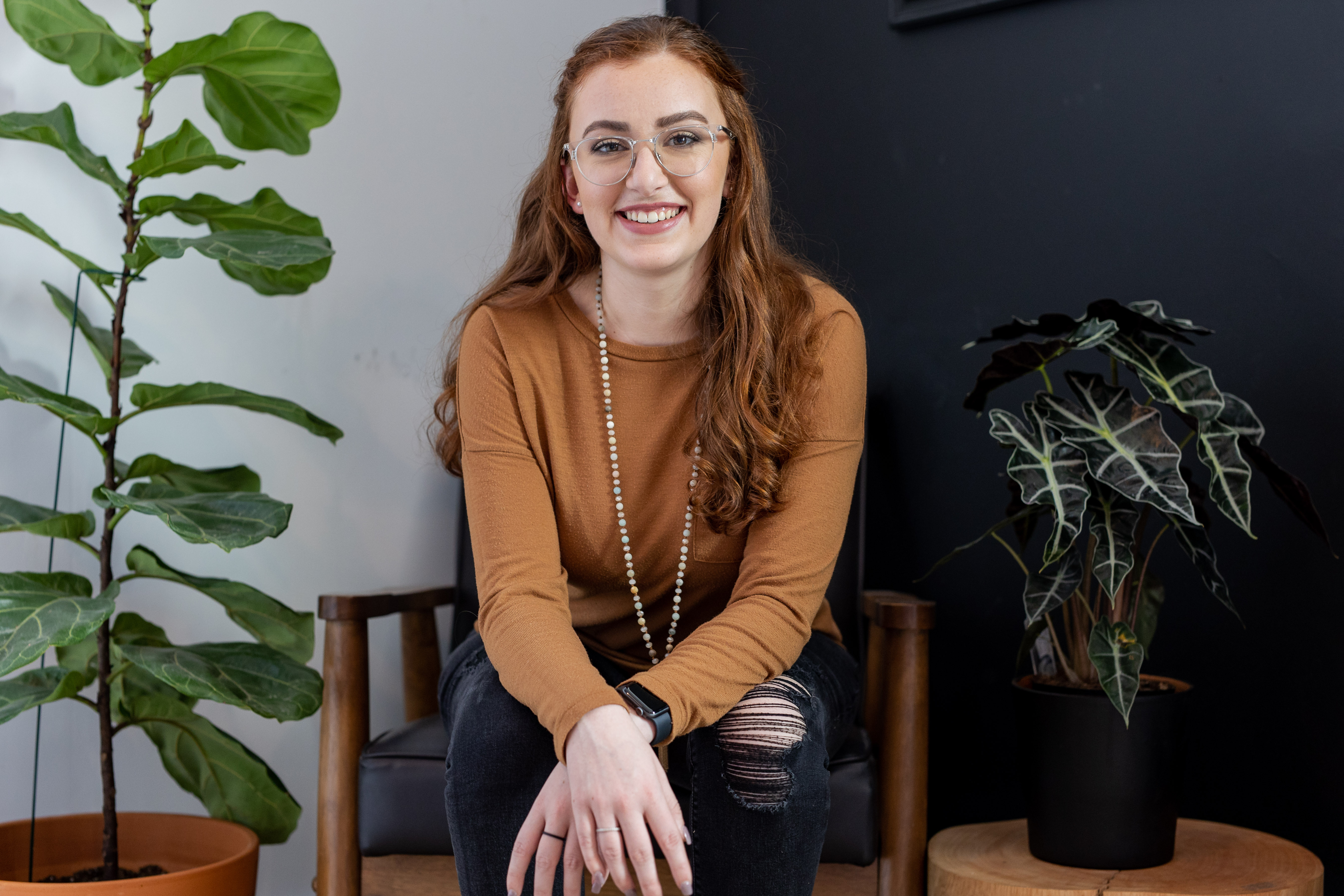 Smiling woman with red hair, glasses, and a brown sweater sitting on a chair between two potted plants.