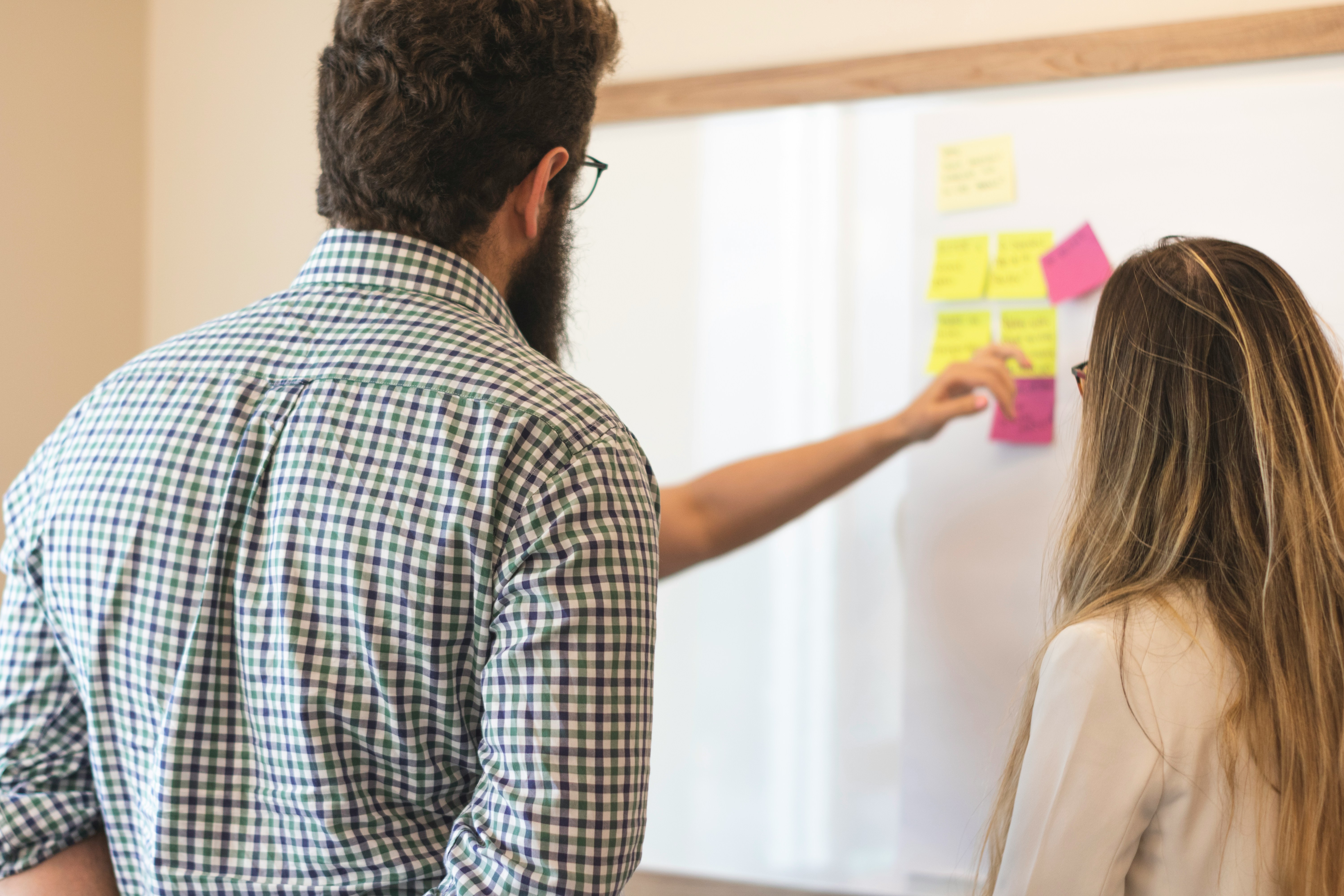 Two coworkers discussing and placing colorful sticky notes on a whiteboard in an office.