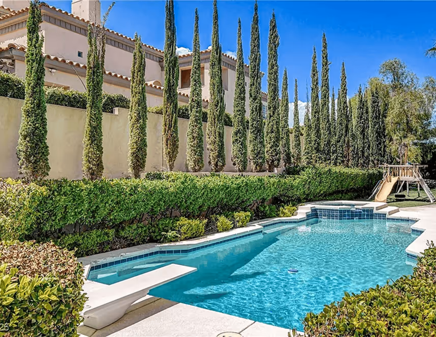 Clear blue swimming pool with a diving board, surrounded by trimmed hedges and tall, narrow trees next to a beige house under a blue sky.