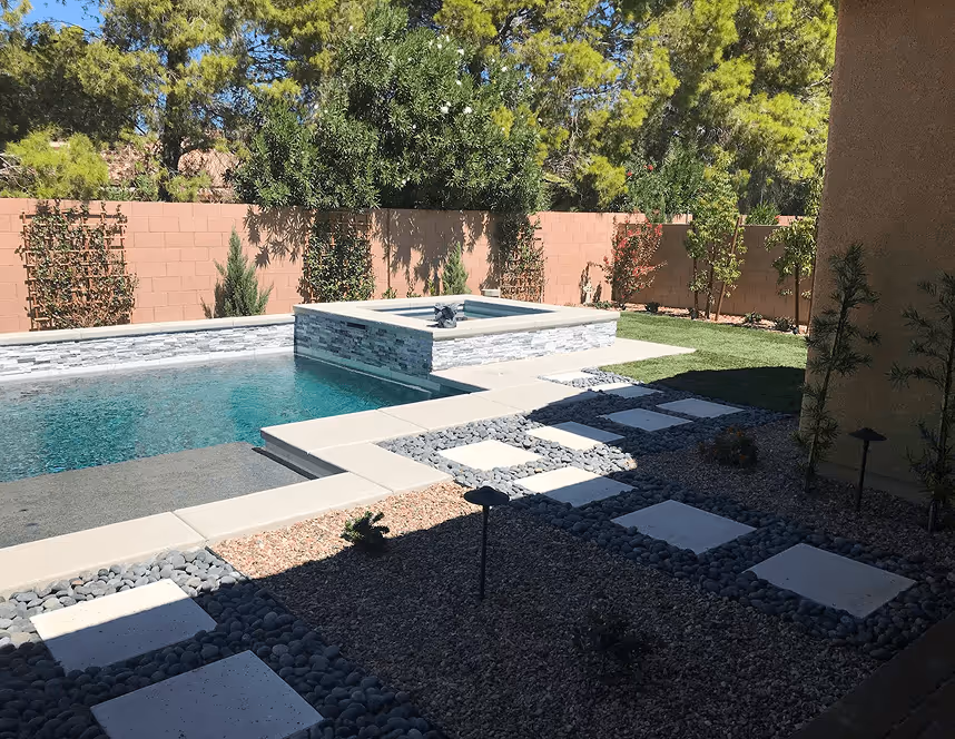 Modern backyard with a rectangular pool, attached raised spa with stone facade, and a pathway of square stepping stones through gravel and landscaping.