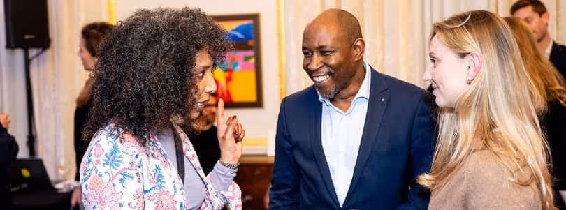 Three people engaged in conversation at an indoor event, with one woman gesturing while the others listen and smile.