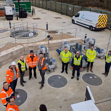 Group of participants of Water Dragons wearing safety vests and helmets standing around industrial equipment outside.
