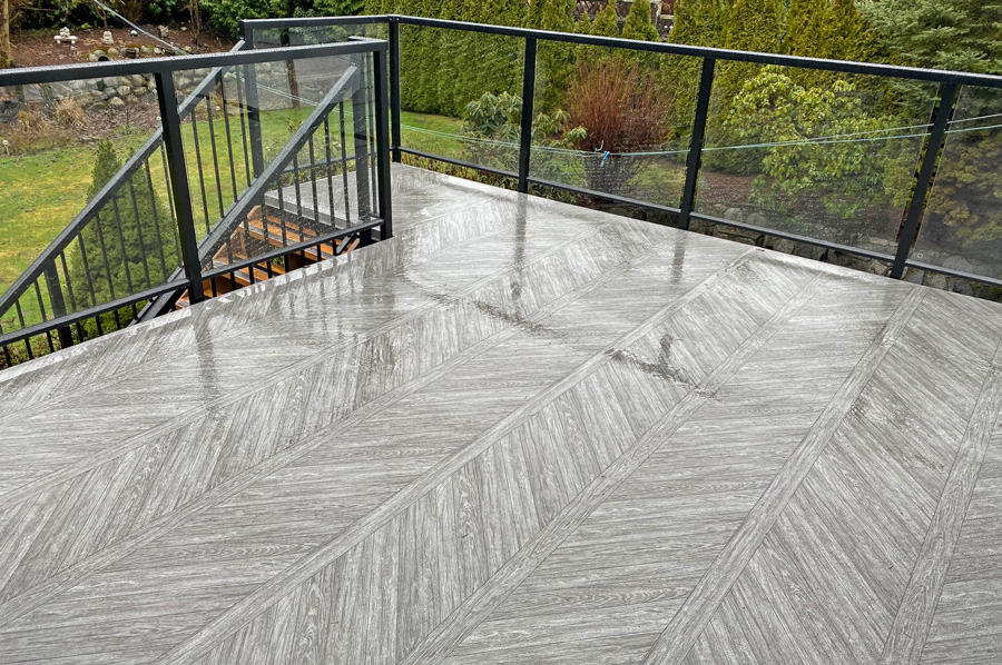 Wet outdoor balcony with gray wood-patterned vinyl flooring and black metal railing overlooking a green garden.