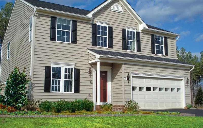 Two-story house with beige vinyl siding, white trim, black shutters, a red front door, and a double garage under a partly cloudy sky.