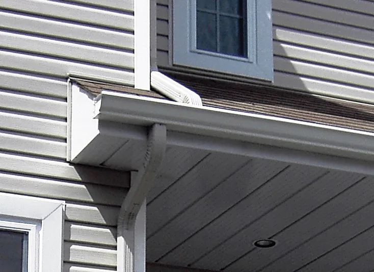 Close-up of a white gutter and downspout attached to a beige siding house with a window above.