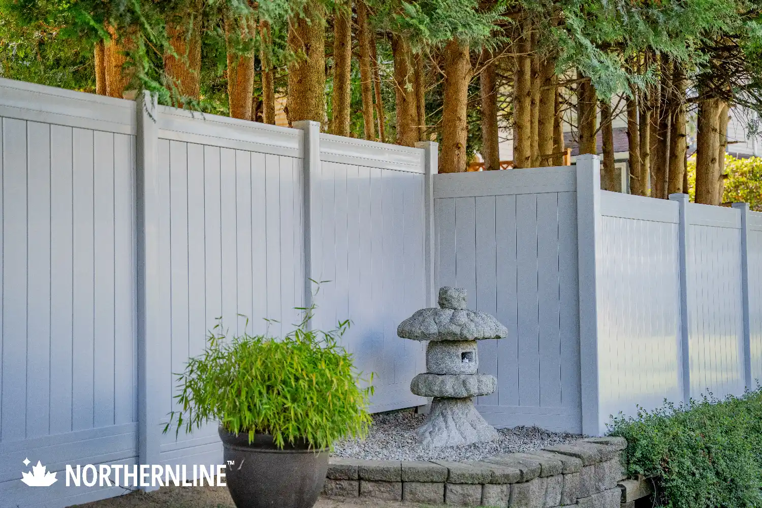 White vinyl fence bordering a garden area with a stone lantern, green plants in a planter, and tall trees in the background.