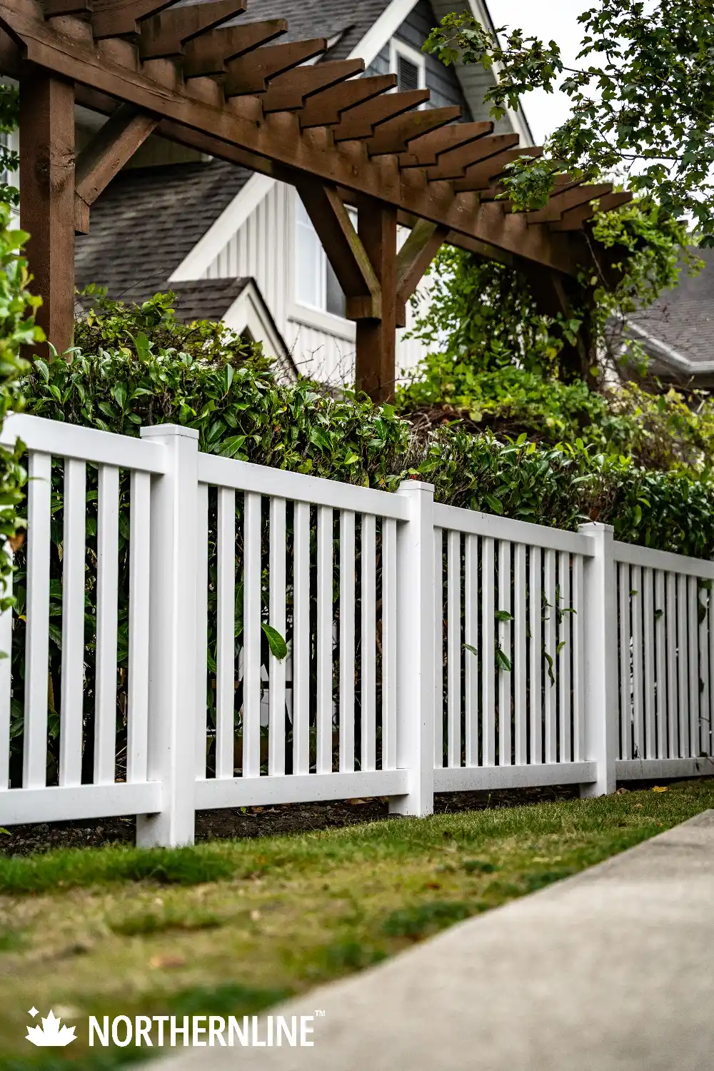 White vertical slat fence with greenery and a wooden pergola in front of a house.