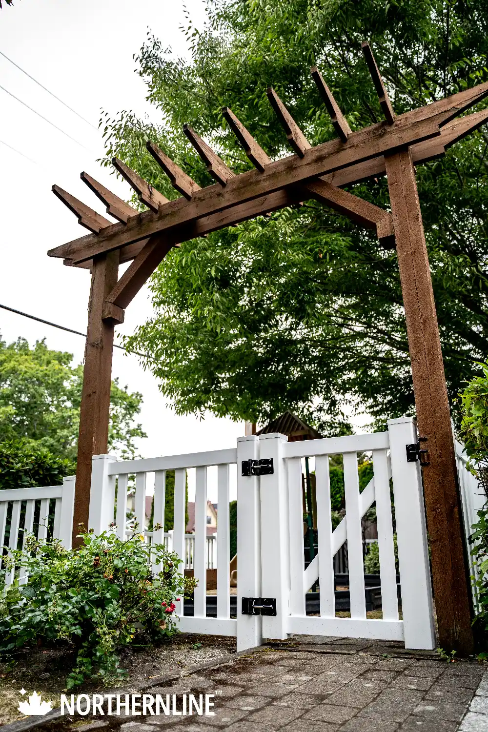 White vinyl garden gate with black hinges under a brown wooden pergola, surrounded by green bushes and trees.