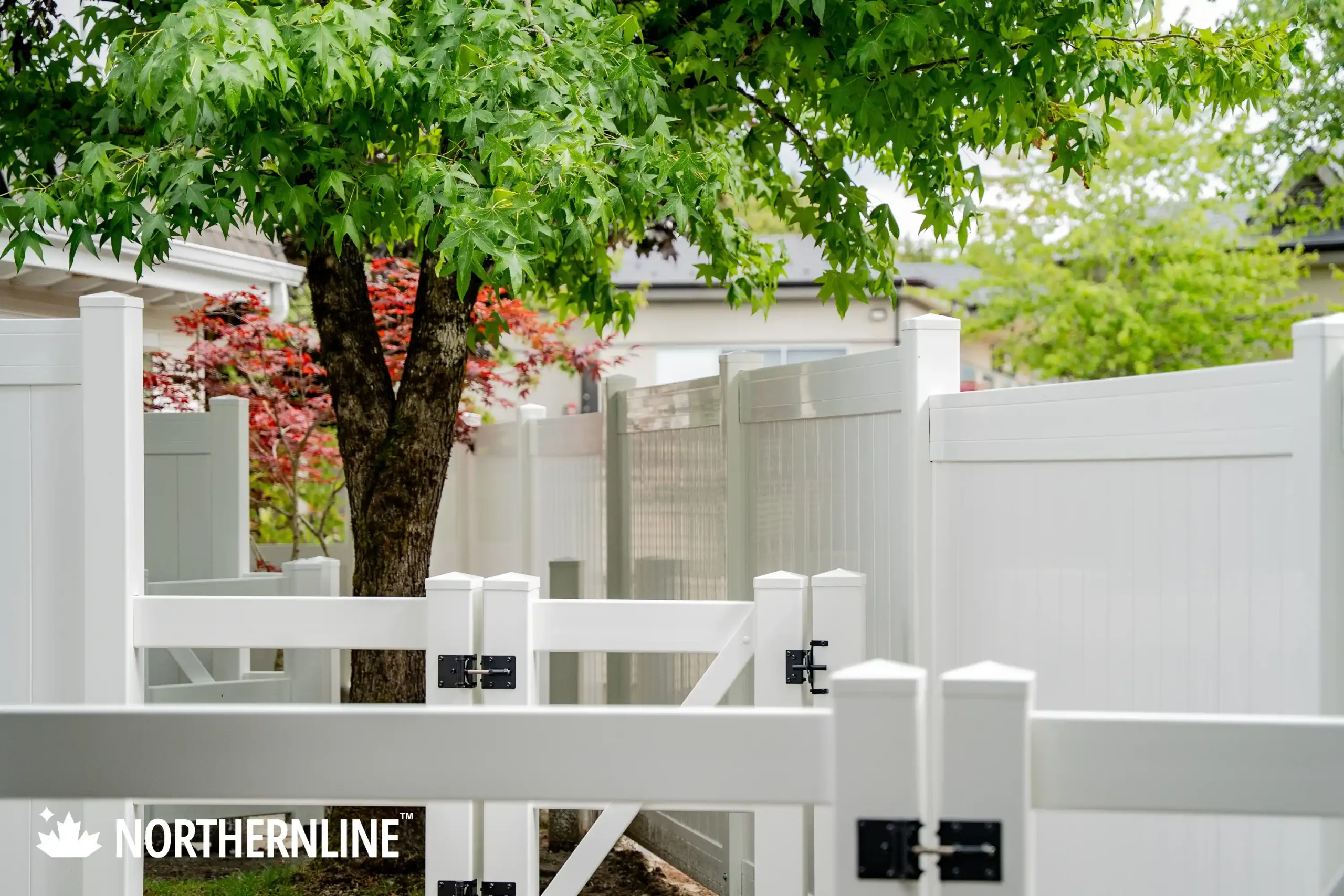 White vinyl privacy fences with gates enclosing a backyard featuring a tree and green foliage.