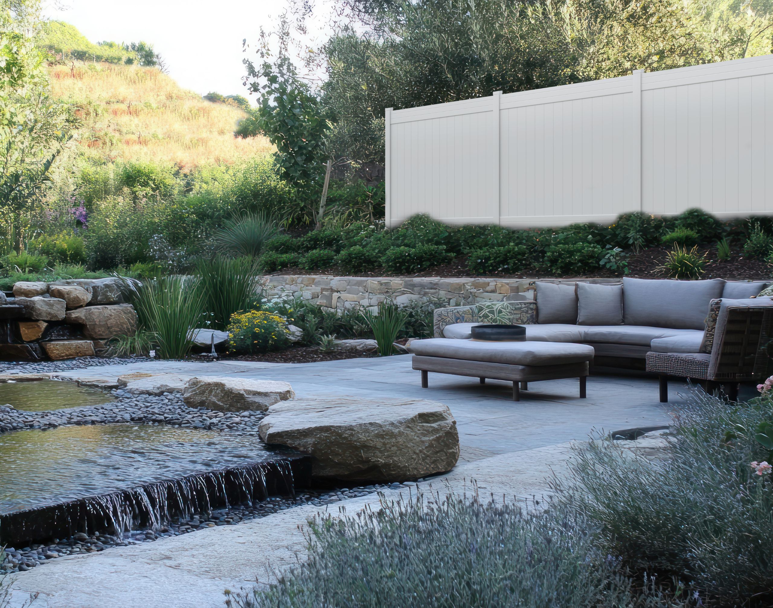 Outdoor patio with gray sectional sofa and ottoman near a stone water feature and lush garden, surrounded by white vinyl privacy fencing.