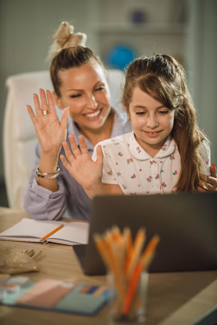 Smiling woman and girl waving at a laptop during a video call at a desk with school supplies.