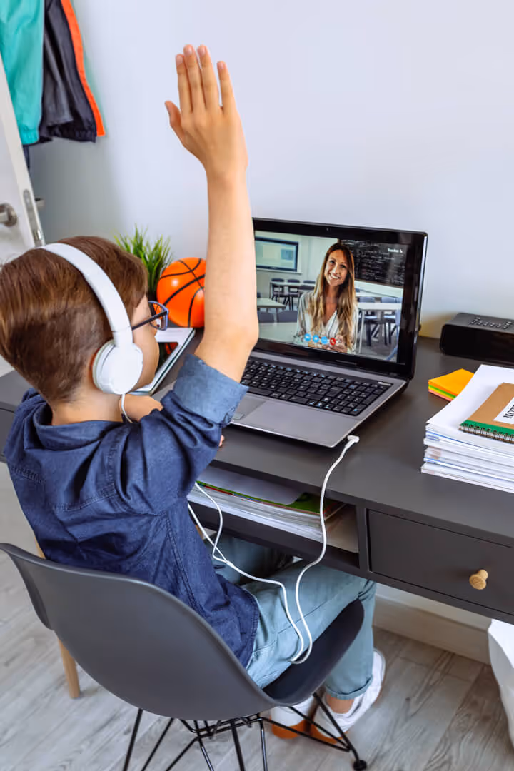 Boy with headphones raises hand during an online class with a female teacher on laptop screen.