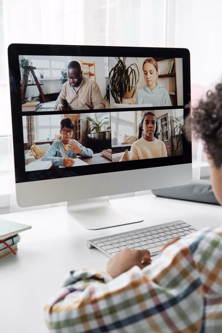 Child sitting at a desk participating in an online video call with four people on a desktop screen.