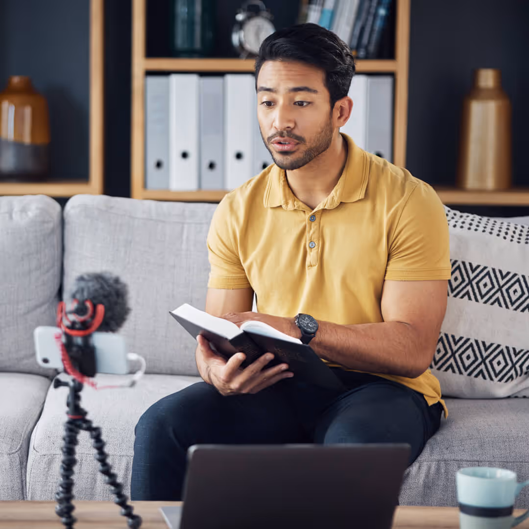Man in yellow shirt reading from book while recording video with phone and laptop on a couch.