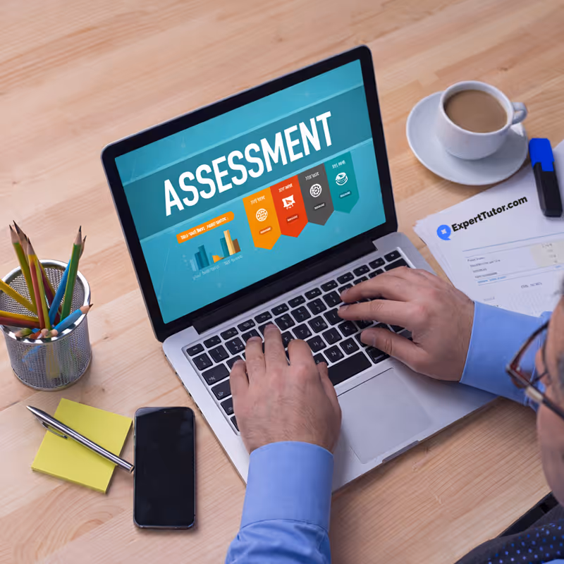 Person typing on laptop with 'ASSESSMENT' on screen, coffee cup, phone, pencils, and paper on desk.
