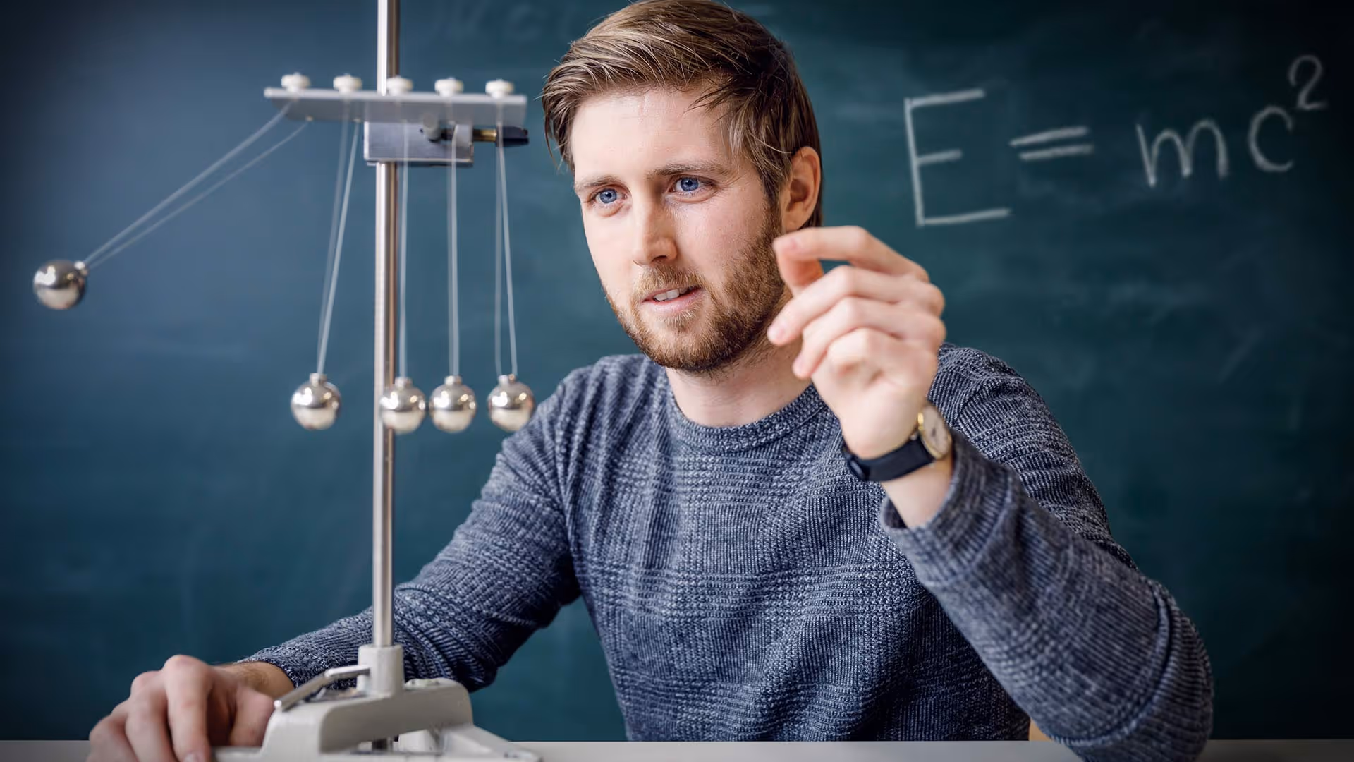 Man demonstrating a Newton's cradle with E=mc² written on a chalkboard behind him.