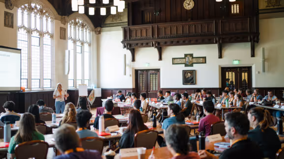 People in a large hall attending a lecture or seminar with a presenter speaking near windows and a screen.