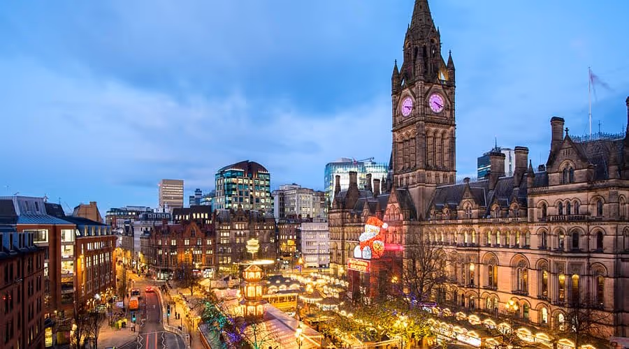 Manchester city hall tower with clock at dusk above a festive market with lights and decorations.