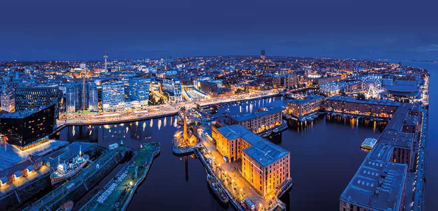 Night aerial view of Liverpool waterfront with illuminated buildings, docks, and a Ferris wheel.