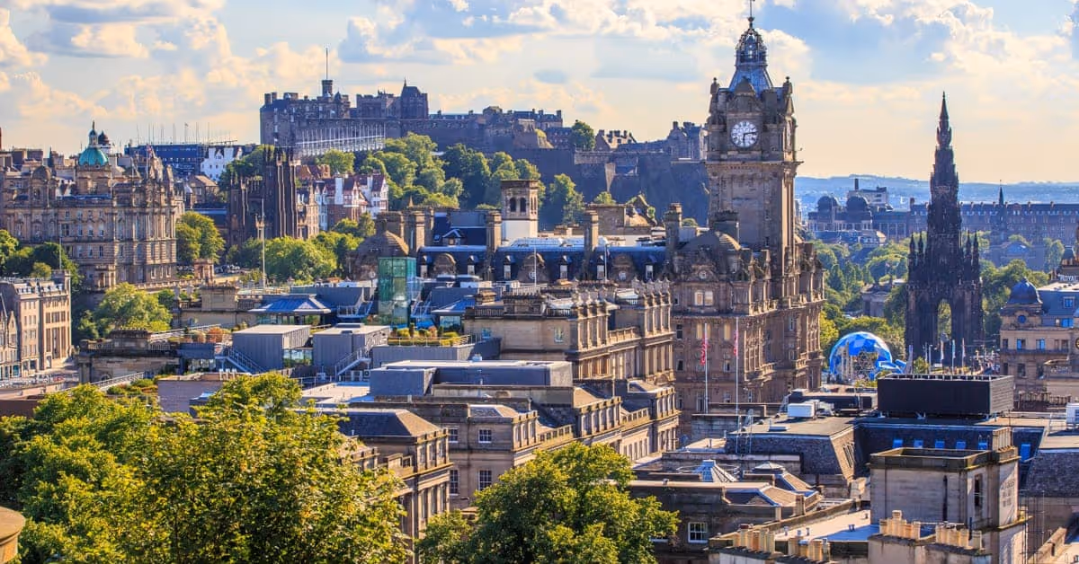 Edinburgh cityscape with historic buildings, clock tower, trees, and cloudy sky in the background.